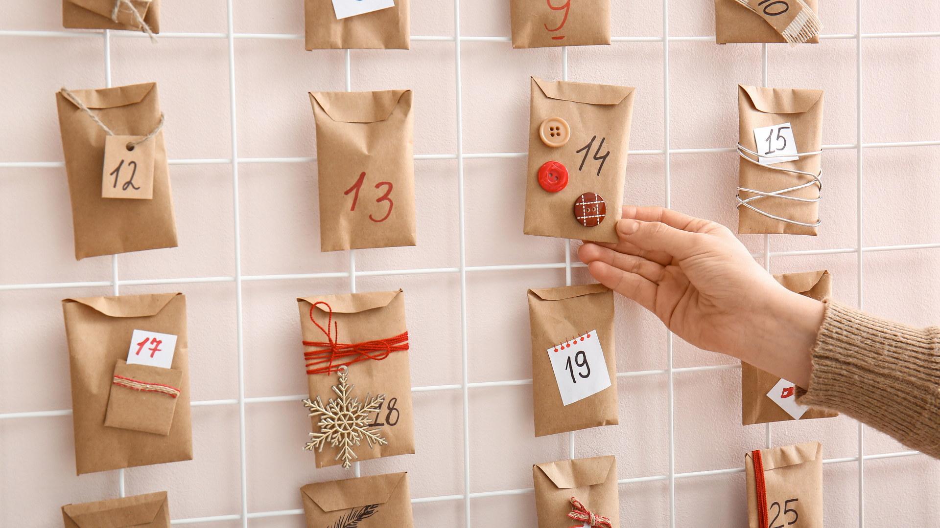 A hand reaches toward a small brown paper envelope labeled “14” on a minimalist DIY advent calendar. The calendar is made of kraft paper envelopes attached to a white metal grid, each decorated with numbers and simple ribbons, buttons, or twine. The background is a soft light pink wall.